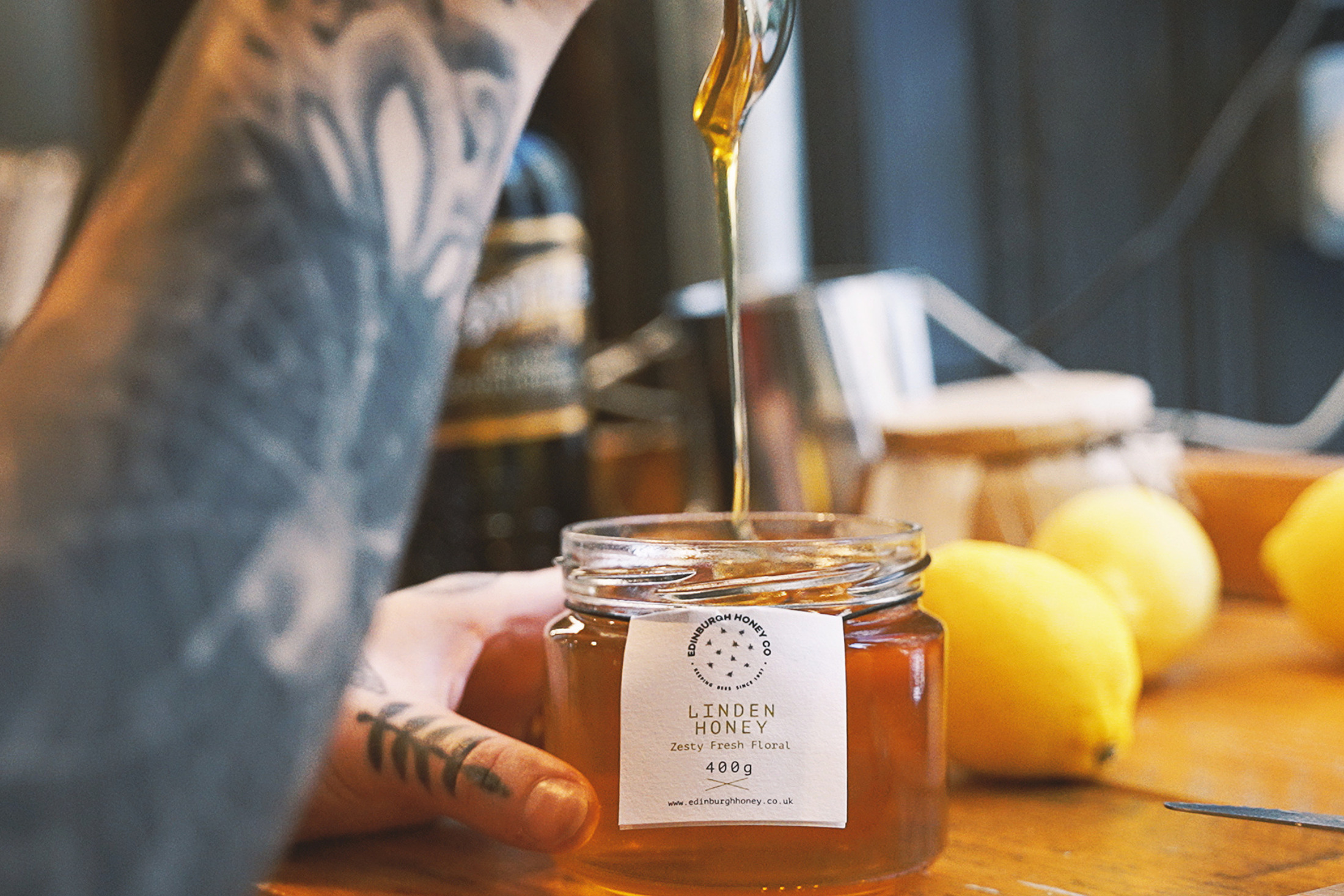 A 400g jar of Edinburgh Honey Co. Linden honey with a tag showing Zesty Fresh Floral a spoon above dripping honey into the jar and lemons in the background