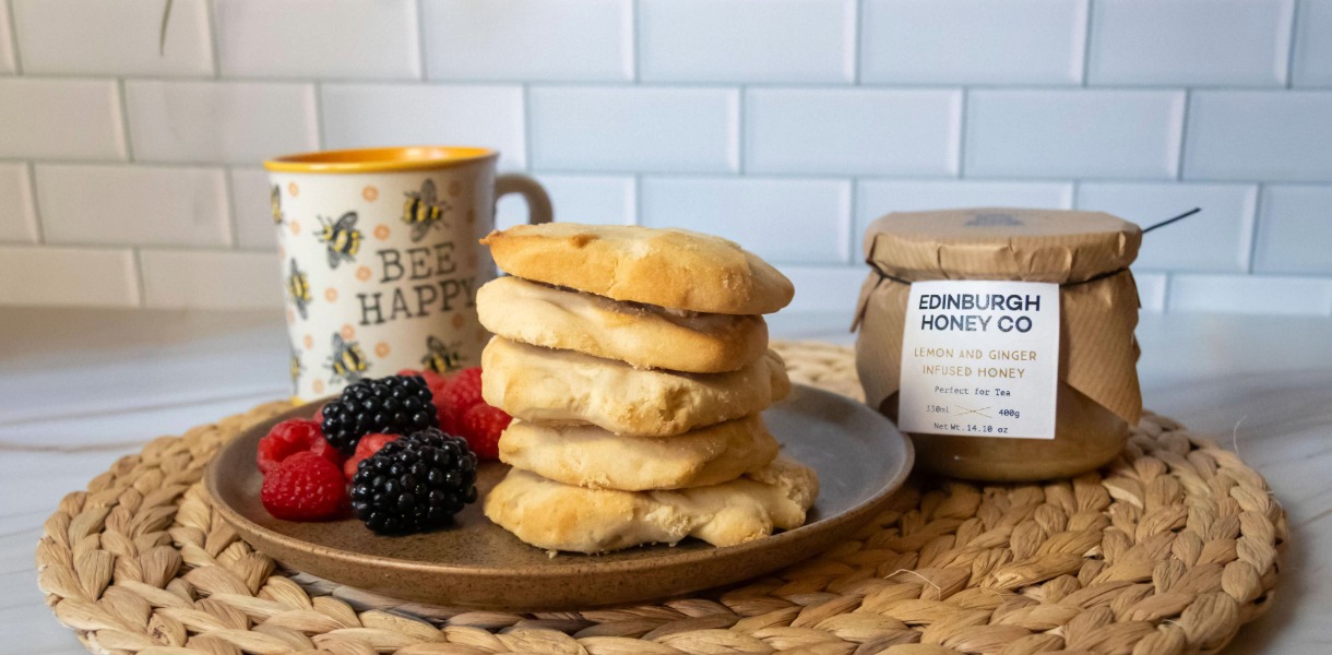 A stack of soft honey shortbread rounds on a ceramic plate with fresh raspberries and blackberries, alongside a jar of Edinburgh Honey Co Lemon and Ginger Infused Honey and a cup of tea